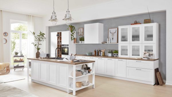 Moderne Küche mit weißen Schränken, einer Insel mit Holzarbeitsplatte, Glasschränken, Pflanzen und Küchenutensilien. Über der Insel hängen Hängeleuchten, und große Fenster lassen natürliches Licht herein. Der Raum ist hell und aufgeräumt.