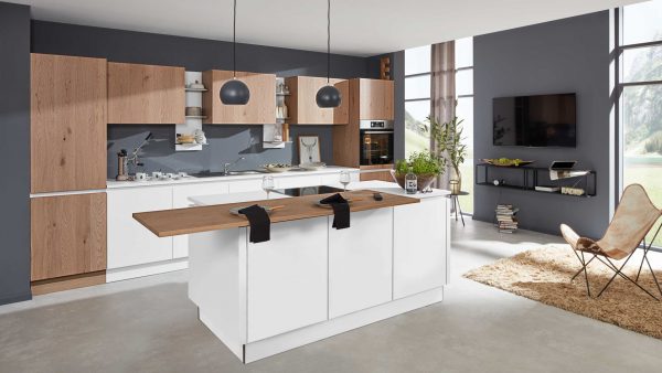 Moderne Küche mit Holz- und weißen Schränken, einer zentralen Insel mit hölzerner Frühstücksbar, zwei schwarzen Hängelampen und einem gemütlichen Wohnbereich mit Fernseher, Sessel und Pflanzen in der Nähe großer Fenster, die viel Tageslicht hereinlassen.