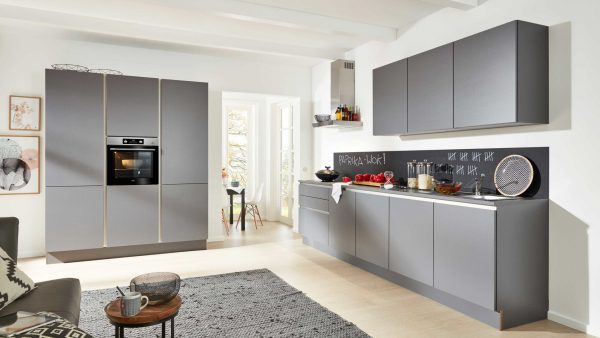 Moderne Küche mit mattgrauen Schränken, Einbaubackofen und schwarzem Backsplash mit einer Kreidetafel mit Schrift und Strichmännchen. Das Sonnenlicht fällt durch Glastüren in einen kleinen Essbereich im Hintergrund.
