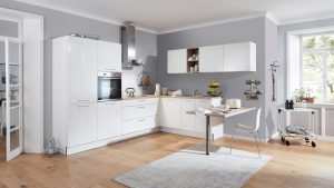 Moderne Küche mit weißen Schränken, Einbaugeräten und einem kleinen Tisch an der Theke. Helle Holzfußböden, graue Wände und ein großes Fenster, das natürliches Licht hereinlässt. Küchenutensilien und Obst sind ordentlich angeordnet.