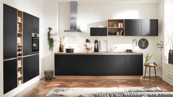 Moderne Küche mit mattschwarzen Schränken, Holzakzenten, eingebautem Ofen und Dunstabzugshaube aus Edelstahl. In den Regalen stehen Geschirr und Pflanzen; an der Wand hängt ein gerahmtes Hirschbild. Sonnenlicht strömt durch große Fenster.