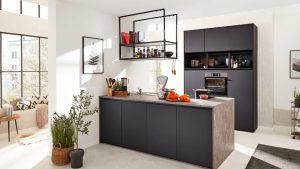 Moderne Küche mit mattschwarzen Schränken, einer grauen Arbeitsinsel, Hängeregalen mit Gläsern und Büchern sowie Küchenutensilien. Sonnenlicht fällt durch große Fenster; auf dem Tresen stehen einige orangefarbene Paprikaschoten und eine Waage.