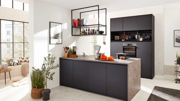Moderne Küche mit mattschwarzen Schränken, einer grauen Arbeitsinsel, Hängeregalen mit Gläsern und Büchern sowie Küchenutensilien. Sonnenlicht fällt durch große Fenster; auf dem Tresen stehen einige orangefarbene Paprikaschoten und eine Waage.