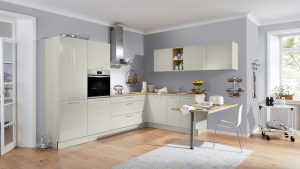 Moderne Küche mit beigen Schränken, Einbaubackofen, Hängeschränken und einem kleinen Tisch mit zwei Hockern. Der helle Holzboden, die grauen Wände und die großen Fenster sorgen für eine helle, luftige Atmosphäre. Küchenutensilien und Kleingeräte sind sichtbar.