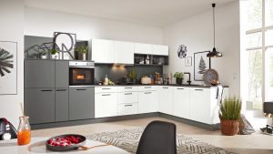 Moderne Küche mit weißen und dunkelgrauen Schränken, Einbaubackofen und verschiedenen Küchengeräten auf der Theke. Der Raum ist hell mit großen Fenstern, Topfpflanzen, Wandkunst und einem gemusterten Teppich auf hellem Holzfußboden.