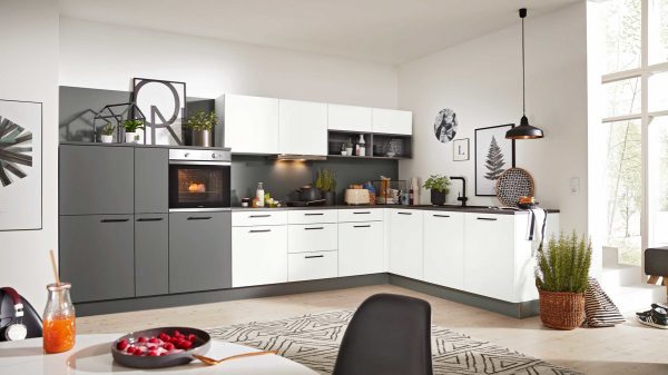 Moderne Küche mit weißen und dunkelgrauen Schränken, Einbaubackofen und verschiedenen Küchengeräten auf der Theke. Der Raum ist hell mit großen Fenstern, Topfpflanzen, Wandkunst und einem gemusterten Teppich auf hellem Holzfußboden.