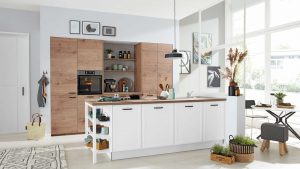 Moderne Küche mit hellen Holzschränken, weißer Insel, offenen Regalen, Backofen und verschiedenen Küchenutensilien. Große Fenster lassen natürliches Licht herein, und Topfpflanzen und Dekoartikel sorgen für eine gemütliche Atmosphäre.