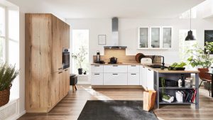 Moderne Küche mit weißen Schränken, Holzakzenten und Geräten aus Edelstahl. Auf der Arbeitsplatte stehen Gemüse und Geschirr, und auf dem Boden liegt eine Papiertüte. Durch die großen Fenster fällt natürliches Licht in den Raum.