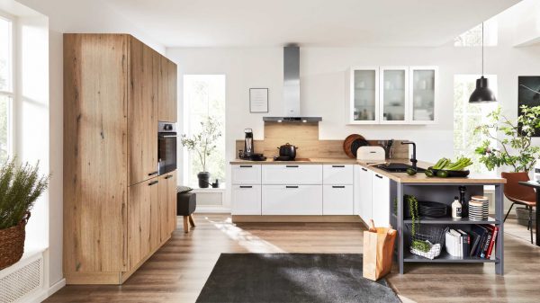 Moderne Küche mit weißen Schränken, Holzakzenten und Geräten aus Edelstahl. Auf der Arbeitsplatte stehen Gemüse und Geschirr, und auf dem Boden liegt eine Papiertüte. Durch die großen Fenster fällt natürliches Licht in den Raum.
