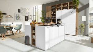 Moderne Küche mit weißen Schränken, Holzakzenten und einer Insel, die mit Geräten und Utensilien ausgestattet ist. In den offenen Regalen stehen Pflanzen und Geschirr; im Hintergrund ist der Essbereich mit grünen Stühlen zu sehen. Große Fenster lassen viel natürliches Licht herein.