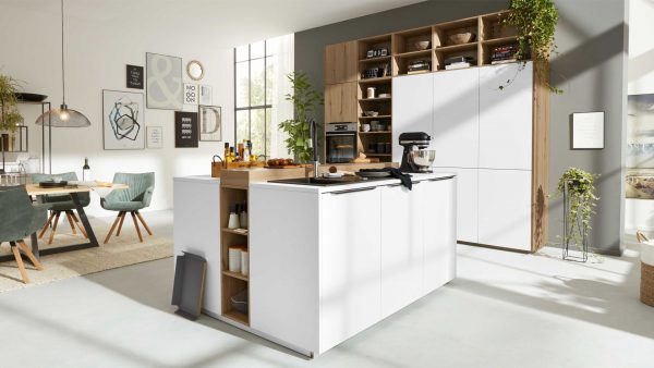 Moderne Küche mit weißen Schränken, Holzakzenten und einer Insel, die mit Geräten und Utensilien ausgestattet ist. In den offenen Regalen stehen Pflanzen und Geschirr; im Hintergrund ist der Essbereich mit grünen Stühlen zu sehen. Große Fenster lassen viel natürliches Licht herein.