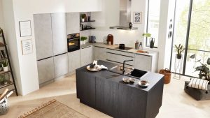 Moderne Küche mit mattgrauen Schränken, eingebautem Backofen und einer schwarzen Insel mit Spüle und Frühstücksplatz. Große Fenster lassen natürliches Licht herein, Pflanzen und Dekoration sorgen für Gemütlichkeit.