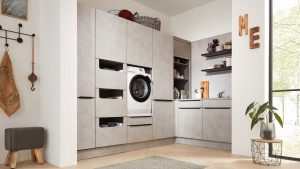 Moderne Waschküche mit schlichten grauen Schränken, einer Frontlader-Waschmaschine, offenen Regalen mit Dekoration, einem kleinen Waschbecken, einer Zimmerpflanze und Sonnenlicht, das durch ein großes Fenster fällt. Das Wort ME ist an der Wand angebracht.