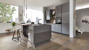 Moderne Küche mit glänzenden grauen Schränken, einer zentralen Insel mit Spüle, einer hölzernen Frühstücksbar, Barhockern, Pendelleuchten und raumhohen Fenstern mit Blick auf die Stadt. Grünpflanzen und Wanddekoration verleihen dem Raum Wärme.