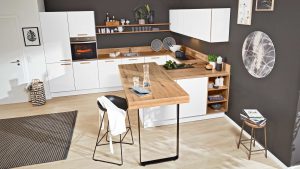 Moderne Küche mit weißen Schränken, Arbeitsplatten aus Holz, schwarzer Akzentwand, offenen Regalen, eingebautem Ofen und einer Holzinsel mit schwarzem Hocker. Dekorationsartikel und Kochbücher sorgen für einen gemütlichen Touch.