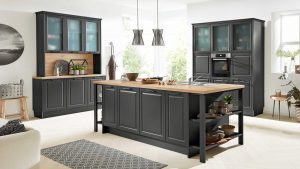 Moderne Küche mit dunkelgrauen Schränken, einer großen Holzinsel mit Regalen, Einbaugeräten, Hängelampen und großen Fenstern, die viel Tageslicht hereinlassen. Dekorationsgegenstände und Küchenutensilien sind ordentlich im Raum verteilt.