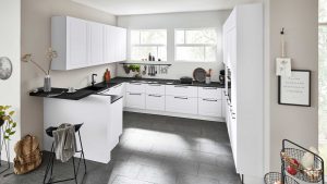 Moderne Küche mit weißen Schränken, schwarzen Arbeitsplatten und schiefergrauen Bodenfliesen. Durch drei Fenster strömt Sonnenlicht herein. Ein Barhocker, ein Korb mit Gegenständen und hängende Dekoration setzen persönliche Akzente.
