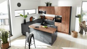 Moderne Küche mit Holz- und Schwarzakzenten, einer Insel mit Holzarbeitsplatte, schwarzem Stuhl, eingebautem Ofen, Spüle, Pflanzen und Küchenutensilien. Große Fenster sorgen für natürliches Licht, und Körbe stehen auf dem Boden.