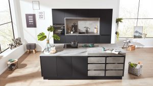 Moderne Küche mit dunklen Schränken, Betonarbeitsplatten und einer zentralen Insel. Zur minimalistischen Einrichtung gehören Pflanzen, Geschirr, eine Teekanne, Wandkunst und große Fenster, die natürliches Licht hereinlassen.