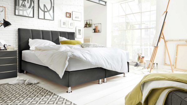 Ein modernes, sonnendurchflutetes Schlafzimmer mit dem Interliving Boxspringbett Serie 1402, einem dunklen Kopfteil und weißer Bettwäsche. Kunstdrucke schmücken eine weiße Backsteinwand, große Fenster lassen natürliches Licht herein und eine gemütliche Dekoration vervollständigt den Raum.
