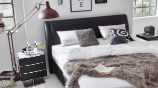 Modernes Schlafzimmer mit dem Interliving Boxspringbett Serie 1401 in schwarzem Leder, weißer Bettwäsche, grauen Kissen und einer grauen Felldecke. In der Nachtkommode stehen Kerzen und Dekoration, auf dem Bett liegt ein offenes Buch. Große Fenster füllen den Raum mit Licht.