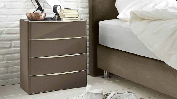 Das Interliving Boxspringbett Serie 1402 - Nachtkommode, ein moderner brauner Nachttisch mit vier Schubladen, steht neben einem Bett mit weißem Bettzeug. Auf ihm liegen Bücher, eine kleine Uhr, eine Metallschale und eine Lampe. Weiße Turnschuhe ruhen auf dem nahen weißen Boden.