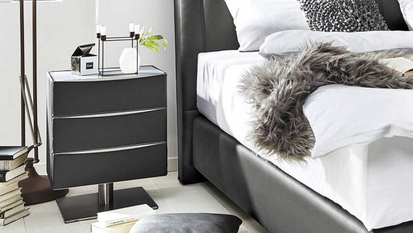 In einem modernen Schlafzimmer steht das Interliving Boxspringbett Serie 1401 - Nachtkommode, ein schwarzer Nachttisch mit drei Schubladen, einer weißen Vase, Büchern und einem Kerzenhalter neben einem Bett mit weißer Bettwäsche, dekorativen Kissen und einer grauen Kunstfelldecke.