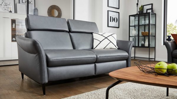 Ein modernes Wohnzimmer mit dem Interliving Sofa Serie 4357 - Dreisitzer 3KV aus glattem grauem Leder, einem weißen Kissen mit schwarzen geometrischen Linien, einem Couchtisch aus Holz mit grünen Äpfeln, gerahmten Kunstwerken und einer Glasvitrine im Hintergrund.