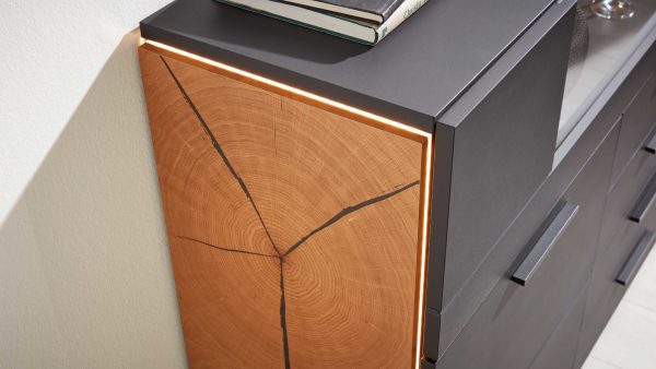 Eine Nahaufnahme der Interliving Wohnzimmer Serie 2023 - LED-Beleuchtung KS502-172 zeigt einen modernen Schrank mit natürlichen Rissen und Ringen in der Holztür, der von einer warmen LED-Beleuchtung hervorgehoben wird, mit Büchern und Dekoration darauf.
