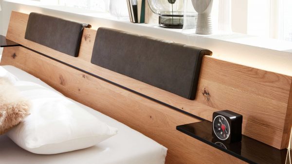 Das Interliving Schlafzimmer Serie 1019 - Kopfteilpolster-Set 523449 verfügt über ein modernes Kopfteil aus Holz, zwei dunkle Kissen, ein weißes Kissen, einen Pelzüberwurf, eine kleine Uhr auf einem schwarzen Regal und dekorative Glasgegenstände auf einer weißen Leiste hinter dem Kopfteil.