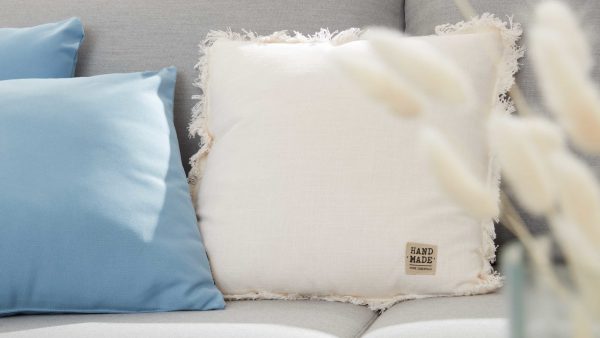 Eine Nahaufnahme der Interliving BEST BUDDYS! Kissen Lenea auf einem Sofa, mit einem blauen Kissen und einem weißen, gefransten HANDGEFERTIGTEN Kissen. Weiche, neutrale Töne und verschwommene Trockenblumen schaffen eine gemütliche, einladende Atmosphäre.