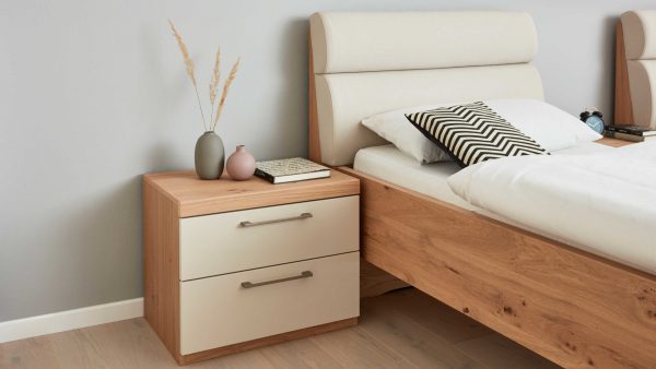 In einem modernen Schlafzimmer stehen ein Holzbett mit weißem, gepolstertem Kopfteil, der Nachttisch Interliving Schlafzimmer Serie 1018 Nachtkommode 220023 mit zwei Schubladen, dekorative Vasen mit getrocknetem Gras, ein rosa Buch und ein schwarz-weiß gemustertes Kissen.