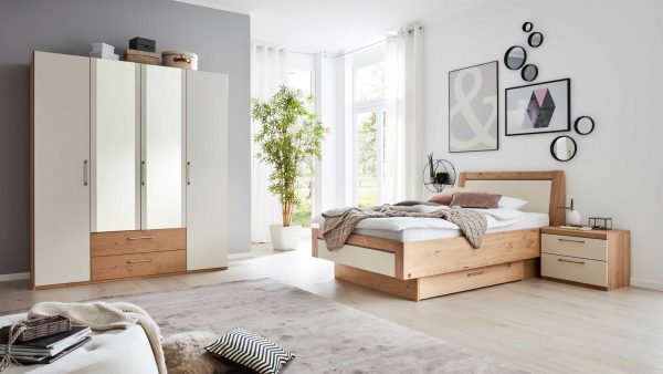 Das Interliving Schlafzimmer Serie 1018 - Komplettzimmer 231 ist mit einem Holzbett, weißem Bettzeug, Nachttisch und Kleiderschrank ausgestattet. Große Fenster lassen viel Licht herein, während Wandkunst und eine Topfpflanze das gemütliche, in neutralen Tönen gehaltene moderne Schlafzimmer aufwerten.