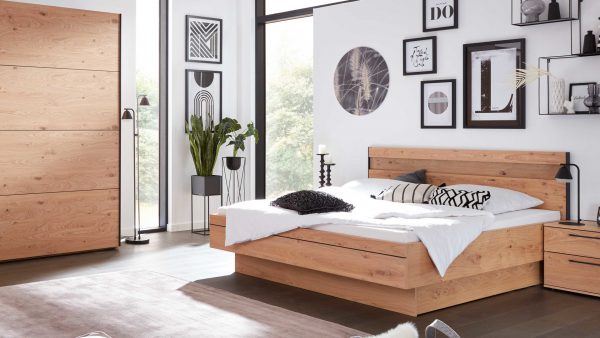 Ein modernes Schlafzimmer mit der Interliving Schlafzimmer Serie 1024 - Bettgestell 86538 in hellem Holz, einem passenden Kleiderschrank, weißer Bettwäsche mit schwarz-weißen Kissen, großen Fenstern, gerahmter Wandkunst und Topfpflanzen in der Ecke.