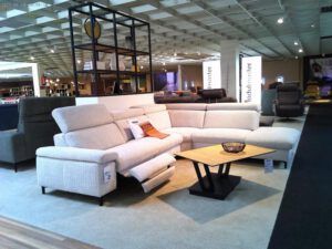 In einem modernen Möbelausstellungsraum steht das Garnitur "Kibo", eine hellbeige L-förmige Couchgarnitur mit eingebautem Ruhesessel, neben einem Couchtisch aus Holz mit einer Drahtschale auf einem hellen Teppich. Im Hintergrund sind weitere Sofas und Displays zu sehen.