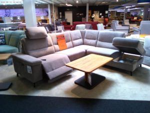 In einem geräumigen Möbelausstellungsraum steht ein großes graues Sektionssofa mit verstellbaren Kopf- und Fußstützen, davor ein hölzerner Couchtisch und im Hintergrund verschiedene andere Sofas und Stühle.
