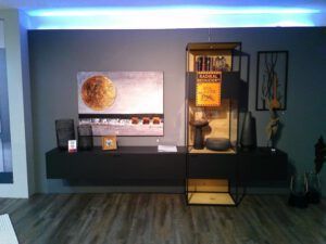 Die Wohnwand "Latoya" besteht aus einem grauen schwebenden Regal, Dekorationsartikeln, Wandkunst mit einem goldenen Kreis und einer hohen Glasvitrine mit einem gelben Verkaufsschild, die vor einer grauen Wand und einem Holzboden steht.