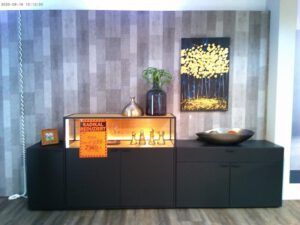 Das Sideboard "Latoya", ein modernes schwarzes Möbelstück mit Vasen, einer Schale und Schachfiguren darauf, steht vor einer grau gemusterten Wand unter einem Gemälde mit gelben Bäumen. Ein orangefarbenes Verkaufsschild steht auf der Anrichte.