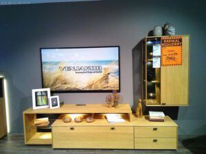 Die Wohnwand "Peking", ein modernes Möbelstück aus hellem Holz mit Schubladen und Einlegeböden, steht unter einem an der Wand montierten Fernseher, der eine Strandszene zeigt. In der Nähe steht ein passender Hochschrank mit einem orangefarbenen Verkaufsschild, beide mit Dekorationsartikeln bestückt.