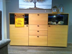 Abgebildet ist das Sideboard "Peking" mit mehreren Schubladen und Glasvitrinen. Ein gelbes Verkaufsschild mit der Aufschrift RADIKAL REDUZIERT kennzeichnet die Preissenkung von 3.515 auf 2.599. Die Dekorationen sind auf der Oberseite angeordnet.