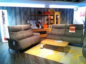 In einem modernen Ausstellungsraum stehen zwei graue Sofas Garnitur IL 4210, ein Couchtisch aus Holz auf einem gelb gemusterten Teppich, Regale mit Dekoration und ein Fernseher, auf dem eine Frau zu sehen ist. Schilder und Etiketten weisen auf den laufenden Möbelverkauf hin.