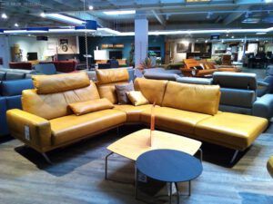 Die gelbe L-förmige Sitzgruppe "IL 4220" von Garnitur mit passenden Kissen steht in einem Ausstellungsraum neben einem Couchtisch aus hellem Holz, auf dem ein kleiner schwarzer Tisch steht. Weitere Sofas sind im Hintergrund zu sehen.