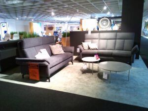 Ein moderner Ausstellungsraum zeigt das Garnitur "Stacey 4" mit zwei grauen Polstersofas, zwei runden Couchtischen und beigem Teppichboden. An den Möbeln befinden sich kleine Schilder, und im Hintergrund sind dekorative Elemente zu sehen.
