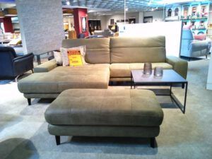 Das grüne Sektionssofa "Dana" von Garnitur mit passender Ottomane und schwarzem Metallbeistelltisch wird in einem Ausstellungsraum zwischen anderen Sofas und Dekorationen ausgestellt, wobei ein Verkaufsschild auf dem Sofa angebracht ist.