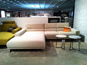 In einem modernen Ausstellungsraum steht die beigefarbene Garnitur "Tossa" mit schwarzen Beinen, zwei runden weißen Nisttischen und Akzentkissen. Eine olivgrüne Couch steht teilweise auf der linken Seite, während rechts eine weiße Backsteinwand zu sehen ist.