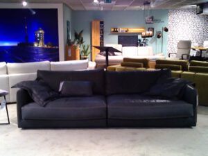 Das Megasofa "Garbo" in schwarzem Leder steht in einem modernen Ausstellungsraum, umgeben von anderen Sofas und Dekorationen, mit einem großen blauen Wandbild im Hintergrund.