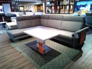 In einem modernen Möbelausstellungsraum steht das große graue Garnitur-Sofa "Serafin" um einen weißen rechteckigen Couchtisch auf einem grün gemusterten Teppich, während im Hintergrund weitere Möbel zu sehen sind.