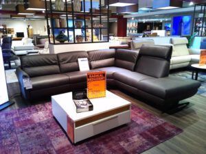 In einem modernen Möbel-Showroom stehen das große dunkelgraue Sektionssofa "Salerno" von Schöner Wohnen und ein weißer Couchtisch mit orangefarbenen Abverkaufsschildern, umgeben von anderen Sofas bei heller Beleuchtung.
