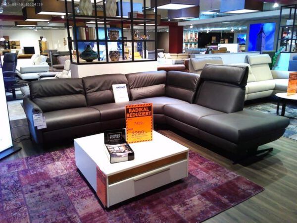 In einem modernen Möbel-Showroom stehen das große dunkelgraue Sektionssofa "Salerno" von Schöner Wohnen und ein weißer Couchtisch mit orangefarbenen Abverkaufsschildern, umgeben von anderen Sofas bei heller Beleuchtung.